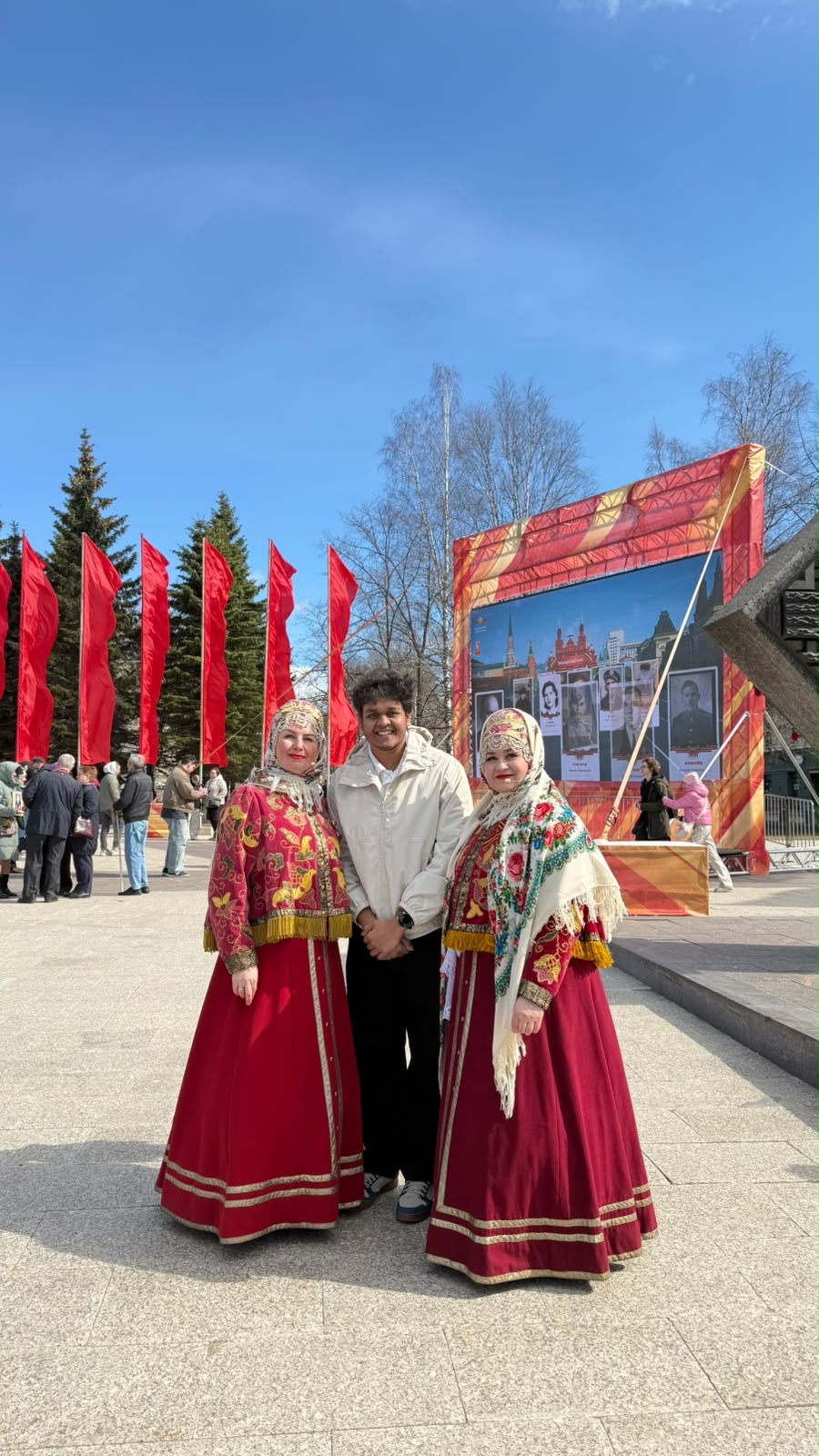 A young man in a cream jacket standing outdoors between two women in traditional red Russian dress, with red flags and trees behind them.